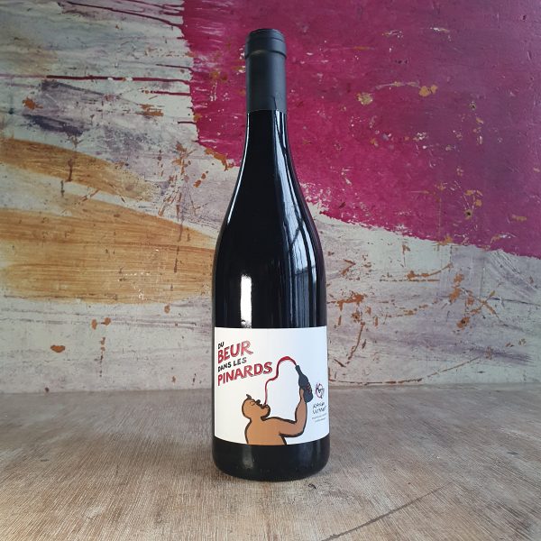 A bottle of red wine on a wooden countertop. The wine's label is white with an illustration of a man drinking wine. The label also features the name 'Du Beur dans les Pinards' and the winemaker's name, Karim Vionnet